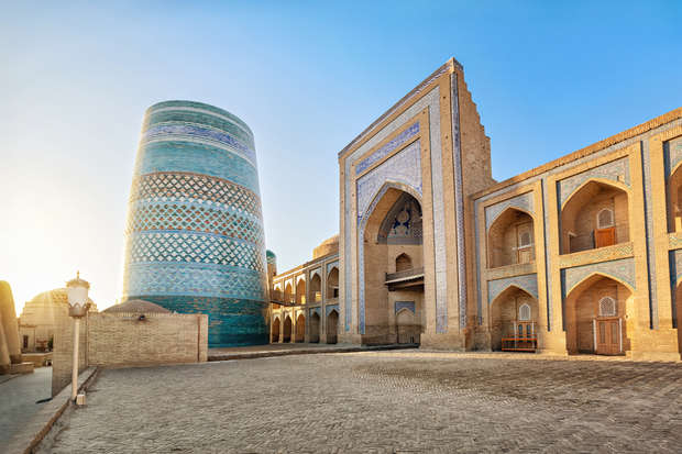 A large, cylindrical tower with teal and white tile patterns stands beside an ornate, arched building. Sunlight casts long shadows on the courtyard, outlining the architectural details.